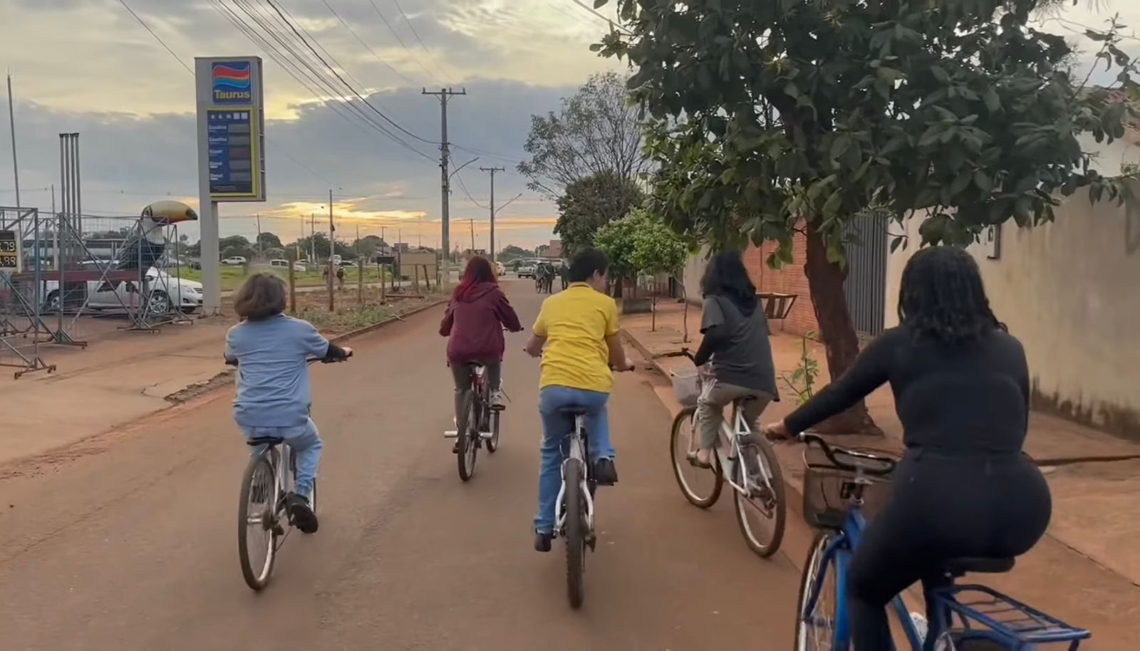 Grupo de cinco pessoas andando de bicicleta em uma rua residencial ao entardecer. Todos estão de costas, pedalando em uma rua arborizada com um posto de gasolina visível à esquerda. O céu está parcialmente nublado, criando uma atmosfera tranquila.